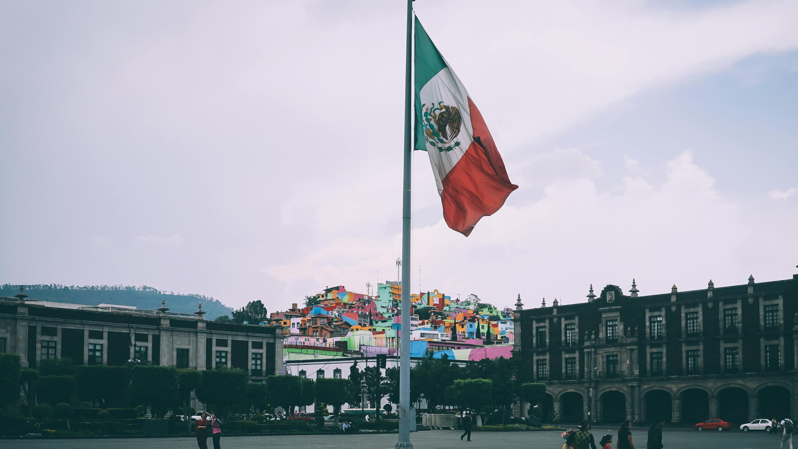 Colorful cityscape of Toluca with large Mexican flag in the foreground, vibrant buildings in the backdrop.